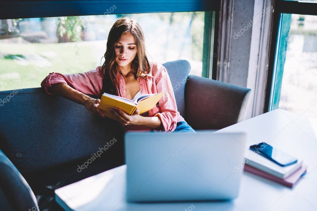 Pensive female student spending free time in cozy cafe reading book and learning information, serious hipster girl analyzing literature while waiting for starting online courses sitting with laptop