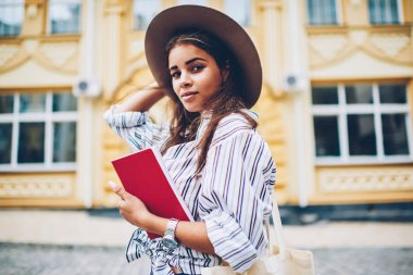 Yarım uzunluk portre çekici öğrenci şık giyim ve moda şapka mimari arka plan üzerinde kamera poz giyinmiş. Kadın edebiyat ile şehir sokaklarında yürürken elinde kitaplar
