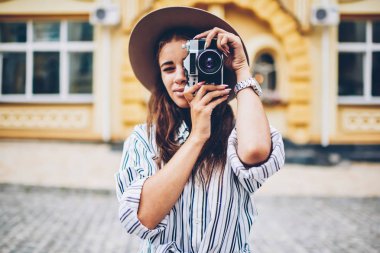 Yarım uzunluk portre odaklanan ve vintage fotoğraf makinesi şehir merkezinin mimari promosyon arka plan üzerinde duran modern lens ile fotoğraf yapma trendy şapkalı şık kadın turist