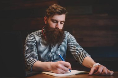 Zeki öğrenci sakal ve ödev alan coworking içinde laptop oturan not defteri olarak aşağı yazma boyun modern kulaklık ile. El denetim listesi kayıt defterinde kalem ile yetenekli genç adam