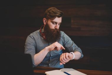 Ev ödevi boşluk dizüstü bilgisayar ve Not Defteri ile coworking oturan zamanında tamamlamak için kol saati göstermek vasıl seyir ve saat yönetme boyun modern kulaklıktan adamla şık hipster sakallı