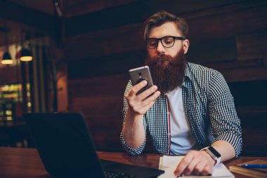 Yüksek hızlı internet bağlantısı kullanarak modern smartphone online bilet rezervasyonu optik gözlük içinde ciddi sakallı metin yazarı. Hipster adam dizüstü cihaz hücresel oturma uygulama yükleme