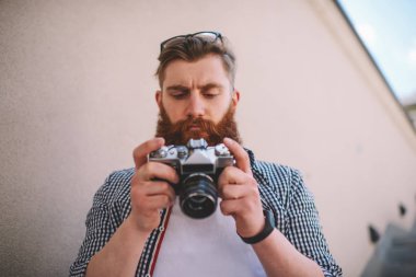Kafasına optik gözlük ile sakallı fotoğrafçı görünümünün altındaki vintage fotoğraf makinesinin ekranda fotoğraf tarama. Işık kontrol ve aygıt açık havada duran açma ayarları yapma rahat tişörtlü genç adam