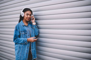 Şık afro Amerikan hippi kız Kulaklığımda favori müzik keyfi denim trendy ceket giymiş Smartphone-e doğru yüklemeleriyle elektronik şarkıları kopyalama alanı alanı yakınında dururken bağlı