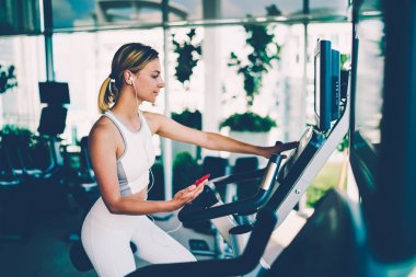 Hız düzey dinleme favori ise eliptik çapraz eğitmen olarak seçerek çekici sportif kadın smartphone cep telefonu ile müzik çalma listesi. Genç kadın eğitim salonunda sağlıklı yaşam destek için uygun