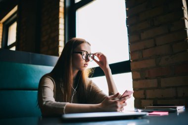 Konsantre yetenekli kız öğrenci telefon eğitim video izlerken gözlük içinde kablosuz internet'e bağlı. Kulaklıklar üzerinde oynarken müzik dinlerken büyüleyici genç manifatura mesaj