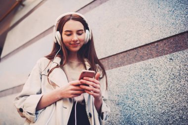 Genç trendy kız öğrenci çalma listesinden müzik dinlemek için uygulama kullanılarak hipster kız açık havada, akıllı telefon ve 4 g Internet bağlantısı kullanarak Web sitelerinde tarama videoları izlerken giyinmiş