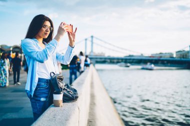 Fotoğrafları kentin güzelliği zevk modern telefonla yapma Eyewear kadın turist. Hippi kız cep telefonu ayakta Köprüsü'nde reklam alanı için reklam yakınındaki nehirde üzerinde fotoğraf çekmek