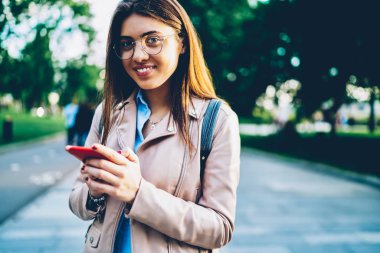 Yarım uzunluk portre çekici gülümseyen Blogger içinde takipçileri ile sohbet ve şehir arka plan üzerinde açık duran kamera bakarken modern smartphone Multimedya paylaşım optik gözlük