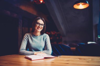 Yarım uzunluk portre oldukça kapalı tahta masada oturan ve ilginç bir kitap okurken kameraya gülümseyen öğrenci akıllı. Gözlük zeki kadın bestseller zevk iyi bir ruh hali var