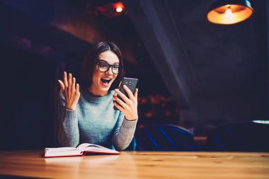 Şık optik gözlük mutlu sürpriz genç kadın Üniversitesi iç eğitim kitabı ile oturan ve bir sonraki sınav için hazırlanıyor smartphone aygıt üzerinde yüksek işaretleri hakkında iyi haber aldı