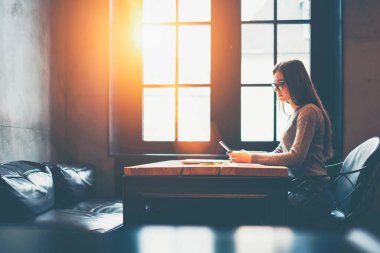 Yan görünüm alanı pencere yakınındaki coworking içinde tahta masada oturan modern smartphone aygıt online ödeme yapma gözlük yetenekli öğrencisi. Genç kadın sosyal ağlar üzerinde hücresel mesajlaşma