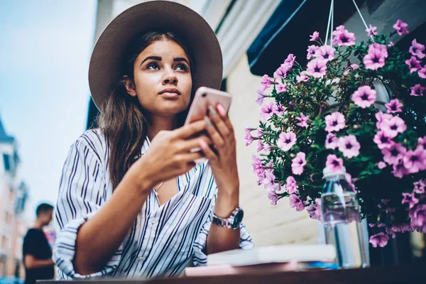 Below view of pondering female travel blogger in stylish hat thinking on publishing internet post of own trip in modern city using telephone sitting at table in cafe outdoors near colorful flowers