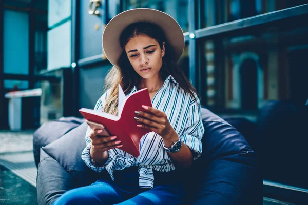 Zeki şık öğrenci eğitim ders kitabı alan coworking içinde yumuşak kabarık oturan ilginç bilgileri okuma trendy şapkalı. Çatı katında oturan kırmızı kitap makale arama yetenekli hippi kız