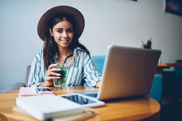 スタイリッシュな帽子の女性ブロガーを浮かべて手にラップトップ コンピューターを持つコーヒー テーブルに座ってカクテルの半分の長さ縦接続ワイヤレス の間にインターネットはカフェでリモートで作業するには — ストック写真