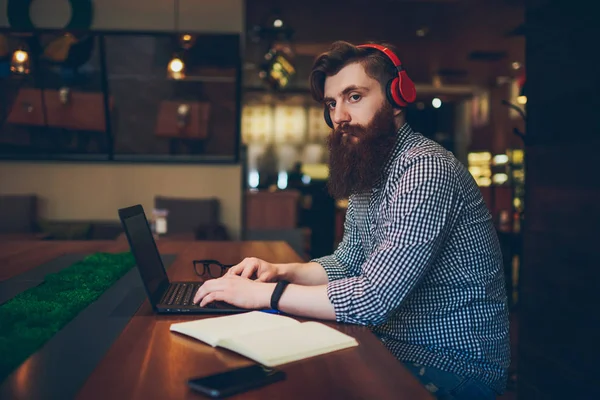 Side view of bearded it professional typing text on keyboard and working freelance at laptop computer connected to wireless internet and listening audio records in headphones sitting in coworking