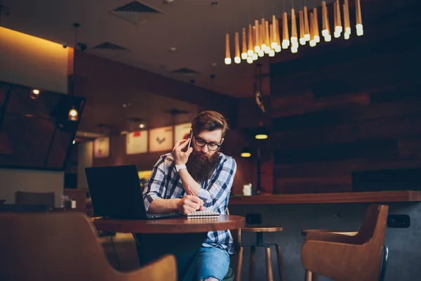 忙碌的年轻胡子男子在眼镜上谈论智能手机设备和写下来的文本在记事本坐在笔记本电脑在咖啡店 时髦的家伙在移动对话中注意到信息 — 图库照片