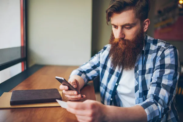 Bearded hipster guy dialing phone number from business card on modern smartphone device sitting in coffee shop.Serious young man dressed in casual shirt sending sms message on digital cellular