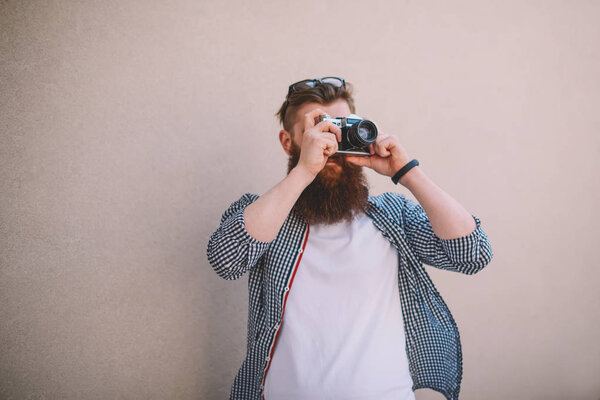 Professional bearded photographer with eyeglasses on head focusing and making photos on vintage camera with modern lens standing on wall with promotional background for your advertising text message