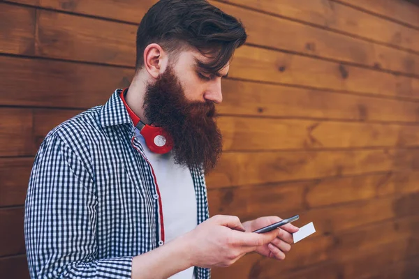 Side view of bearded meloman with stylish red headphones on neck dialing telephone number from business card on modern smartphone standing outdoors.Wooden promotional background with copy space area