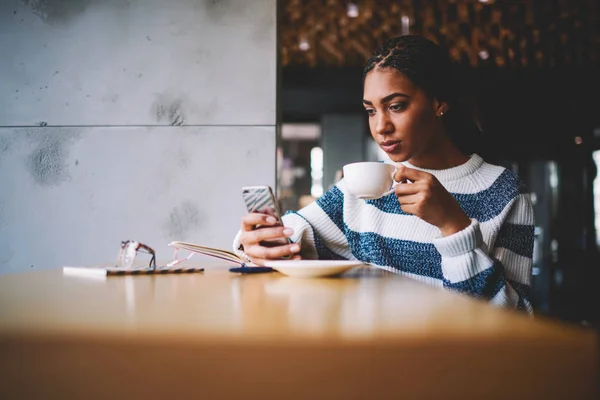 深刻なアフロ アメリカ若い女性手に一杯のおいしいコーヒーとコーヒー ショップのインテリアで休んでいる高速のインターネット接続を介して近代的なスマート フォンで Web 店ショッピングを行います 宣伝エリア — ストック写真