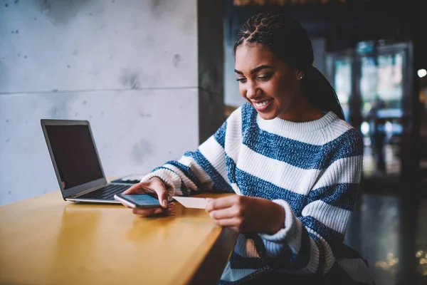 Happy young woman looking at business card and dialing phone number on smartphone for future job on internet website.Positive copywriter with telephone in hand sitting at laptop computer