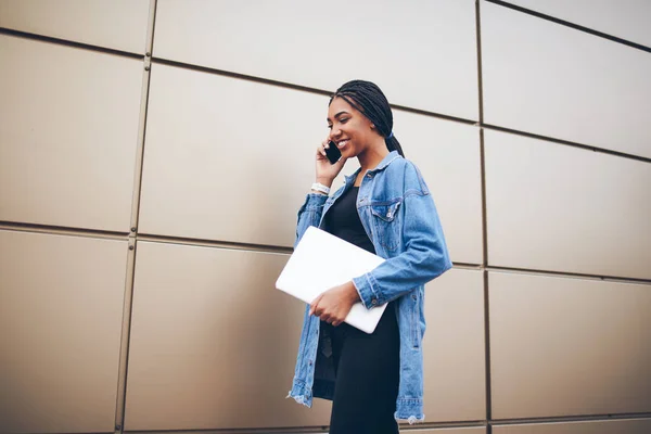 Cheerful female blogger dressed in trendy apparel with dark skin communicating with friends about successful project on telephone strolling along promotional background with laptop computer in hands