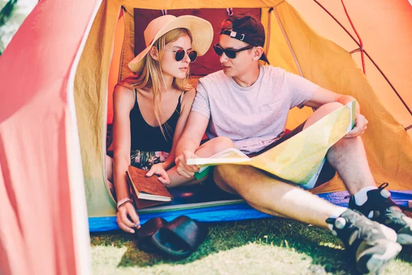 Male traveler talking with girlfriend making plan for journey reading map resting in tent,romantic hipster couple recreating together camping during summer vacation camping discussing next route