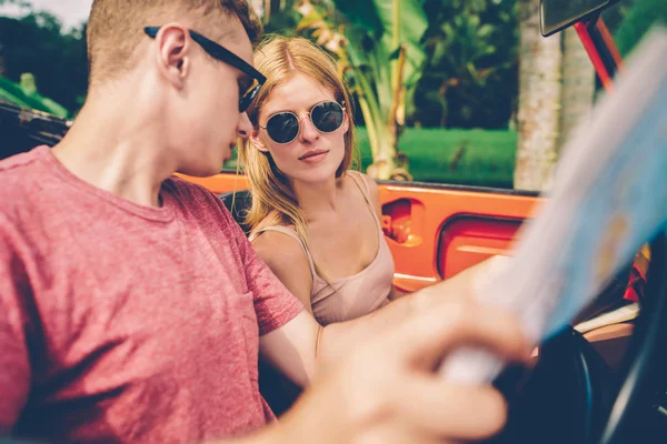 Couple in love discussing ideas for route reading map while sitting in rental vantage car,male and female hipsters driving on cabriolet exploring tropical island during summer vacations together