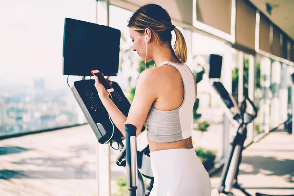 Woman watching video about training in gym on cellphone connected to internet while making exercises on bike.Young fit female selecting motivational music composition during fitness workout in gym