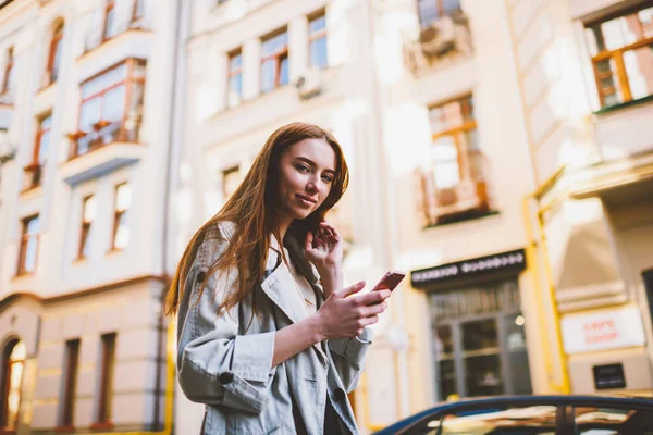 Portrait of positive red hair woman looking at camera while holding gadget in hand.Beautiful  tourist walking at street and sharing impressions with friends in social networks on smartphone
