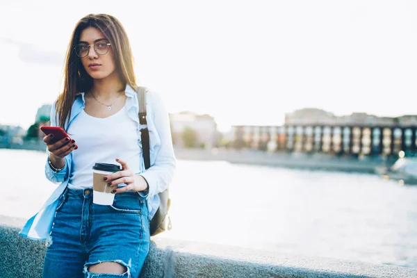 有吸引力的黑发旅行者的画像在眼镜检查电子邮件在现代 Smarthone 站立与咖啡在手外面 女性游客发送短信时 看着镜头附近的宣传区域 — 图库照片