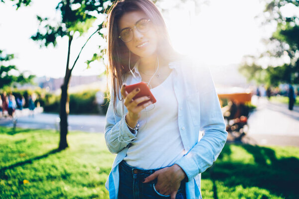 Полудлинный портрет красивой веселой молодой женщины, слушающей электронную музыку из плейлиста в смартфонах, подключенных к смартфону, стоящим на открытом воздухе в парке с солнечными лучами на зеленом фоне
