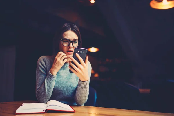 Hipster girl in eyeglasses worrying and watching online broadcasting on mobile phone while preparing for exam.Emotional female reading student\'s rating on university website in expecting good result
