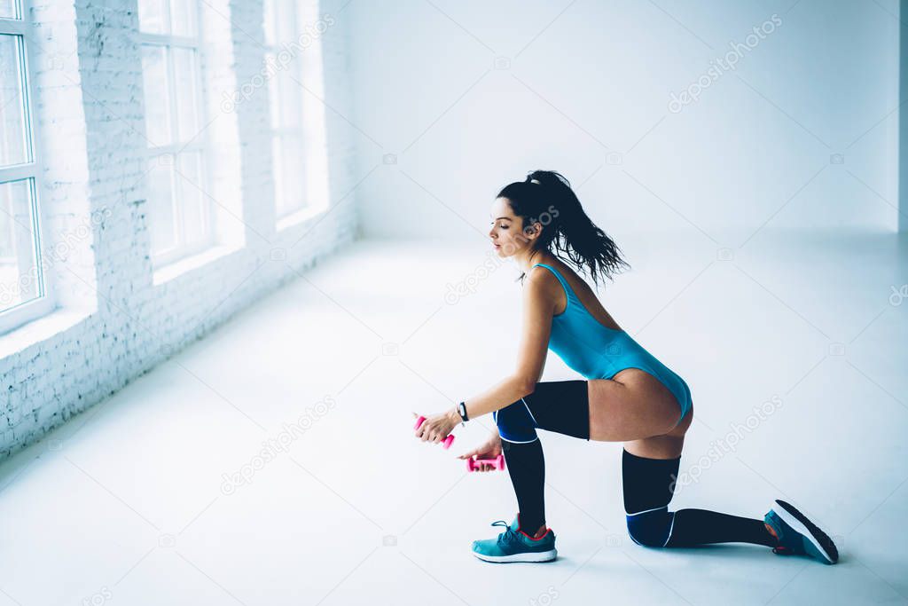 Young sportswoman in tracking suit using dumbbells for squatting training lower body,sexy fitness girl having hard workout doing aerobic exercises for keeping perfect body shape reaching goals
