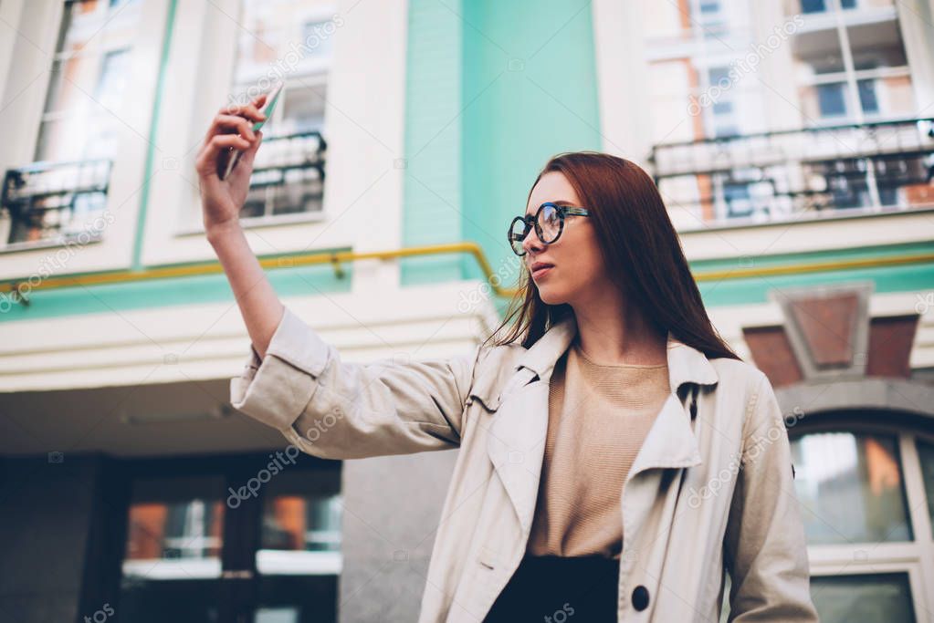 Pensive charming hipster girl making selfie on smartphone camera sharing photo in social networks while scrolling on spring day, female tourist in trendy wear making picture on urban setting background