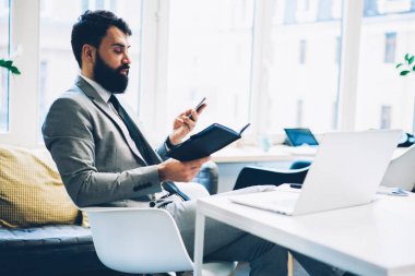 Sakallı erkek bankacı modern smarthone reklam mesajı için netbook ofis iç tanıtım alanının yakınında oturan mobil uygulama yüklenirken çalışma shedule siyah Not Defteri'nde okuma