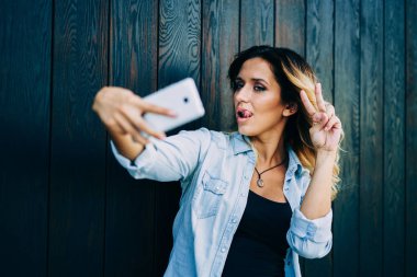 Genç çekici blogger selfie yapım ve barış gösterilen süre hücresel kamera için poz. Fotoğraf çekme ve alan reklam yakın duran dudakları dil koyarak sırasında dalga serin kız öğrenci