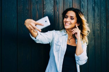 Neşeli kadın blogger hücresel kameraya selfie yaparken poz şık kıyafeti giymiş. Oldukça olumlu öğrenci dinlenme sırasında ayakta açık havada yanında kopya alanı ile duvar zevk