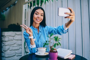 Güzel neşeli kadın kişi barış selfie modern telefon kamera alarak sırasında gösterilen yarım uzunluk portre. Şirin başarılı esmer blogger cep üzerinde selfie yaparken kameraya bakıyor