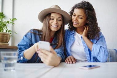 Smartphone ve sosyal ağlar hücresel zevk ücretsiz zamanında birlikte komik video izlerken neşeli hippi kız kafe, dinlenme paylaşım fotoğrafları inceleyen çok ırklı arkadaşlar gülümseyen