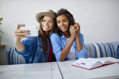 Neşeli çok ırklı hipster kızlarda Afro-Amerikan arkadaşı ağlar üzerinden cellphon payı için kadın alarak resim gülümseyen trendy kıyafetler selfie kafe, smartphone kamera için poz