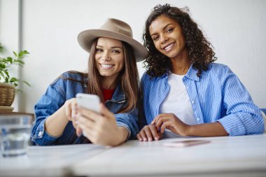 Birlikte eğleniyor neşeli hippi kız portresi bloglama sosyal ağlar için smartphone kullanarak, rezervasyon yapma kafede oturan kamera bakarak genç en iyi arkadaşlar web sayfası