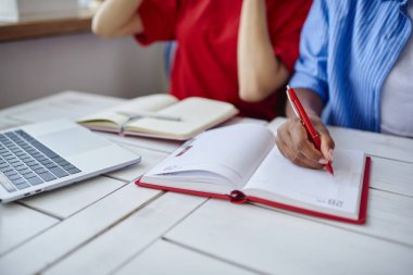 Çok ırklı kız öğrencilerin ödev görev, kadın eli kalem yapılacaklar listesi oluşturma ve proje içinde not etmek ahşap deskto oturan için planlama yaparken Not Defteri'nde yazma kırpılmış görüntü