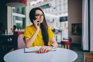 Esmer kadın mali Yöneticisi'nde modern cep telefonu aygıtta meslektaşı ile konuşmak ve uzak kapalı kafeteryada oturan bakarken eserin konuları tartışmaya şık optik gözlük