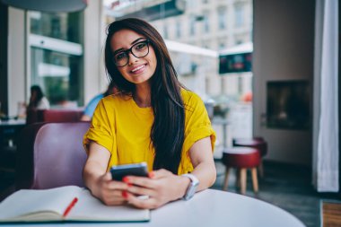 Yarım uzunluk portre neşeli genç girişimci hızlı 4 g internet bağlantısı üzerinden telefon aygıtındaki arkadaşına online transfer para yaparken kameraya gülümseyen optik gözlük