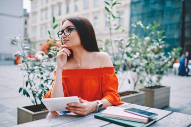 Esmer öğrenci uzak webstore moda giyim kentsel ortamda kitap ve tablet ellerde ile masada oturan oluşturma iş planı üzerinde düşünürken seyir gözlük rüya