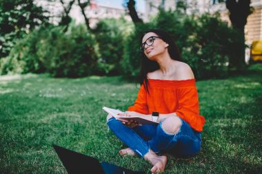 Optik Gözlük üzerinde düşünme rüya yetenekli öğrenci kitap açılan dizüstü bilgisayar ile yeşil çimenlerin üzerinde oturan bölümünü okuyun. Hippi kız zevk bestseller. Reklam alanı için reklam