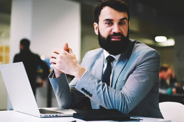Portrait of cheerful bearded financial director dressed in formal clothing smiling at camera while working at modern laptop computer device connected to wireless 4G internet in office building