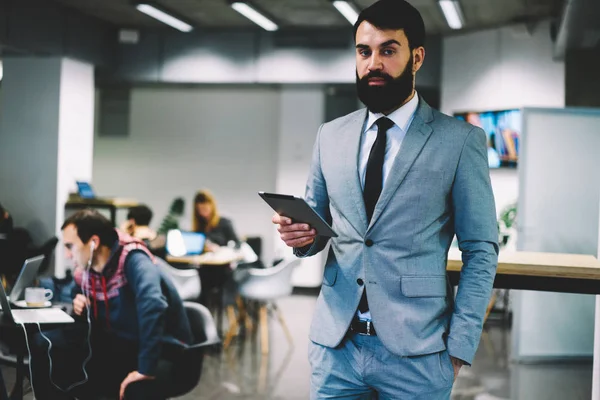 Portrait of stylish corporate director in formal clothing looking at camera while checking email on digital tablet connected to free 4G internet standing indoors in office with professional employees